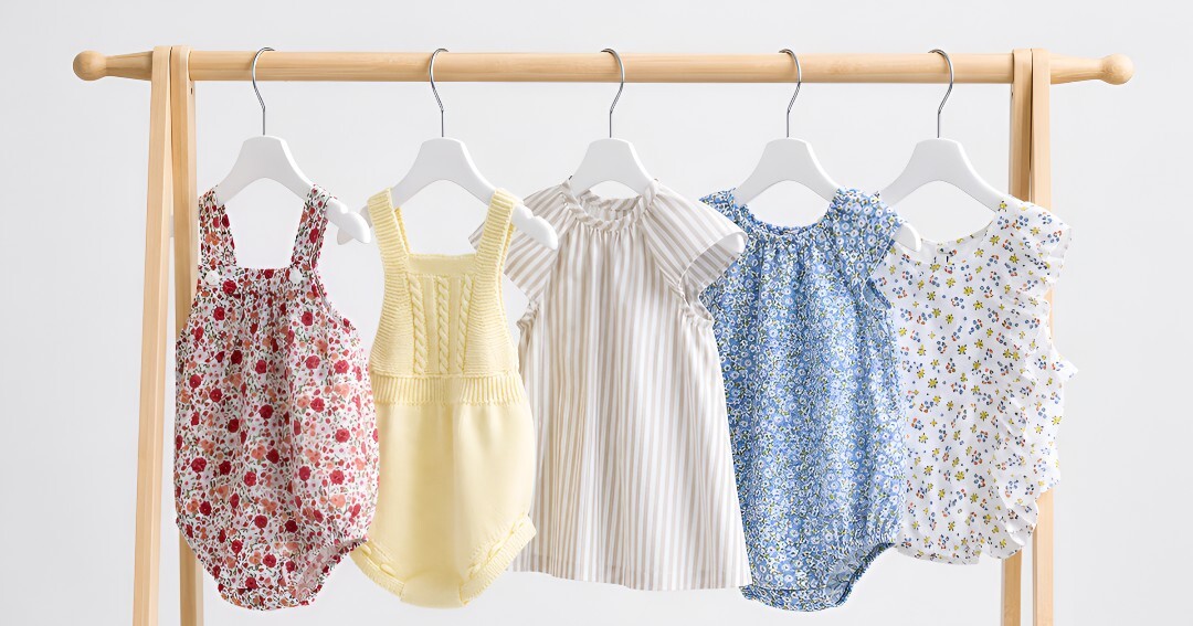 Un appendiabiti in legno su cui sono esposti i capi estivi per neonati della collezione Babydif, tra cui tutine in cotone e indumenti leggeri e traspiranti – perfetti da mettere in valigia per le vacanze.